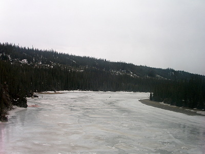 [The lake looks like a wide river which disappears around the bend to the right behind all the evergreens which actually line both sides of the ice. There appear to be a few small patches of water, but it may just be thinner patches of ice. The sky is completely cloud-covered so the image nearly appears to be a black and white one even though it is in color.]